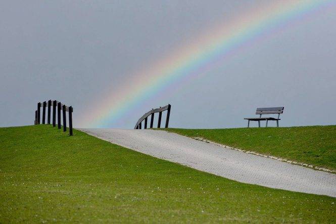 Ein Beeindruckender Regenbogen Erstreckt Sich über Den Grünen Deich, Während Eine Einsame Bank Darauf Wartet, Dass Besucher Den Malerischen Ausblick Genießen.
