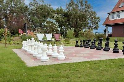 The Large Garden Chessboard Is Surrounded By Lush Green Lawns And Colorful Flower Beds. The Life-size Chess Pieces Are Placed On The Tiles Of The Chessboard, Waiting To Be Moved In An Exciting Game.