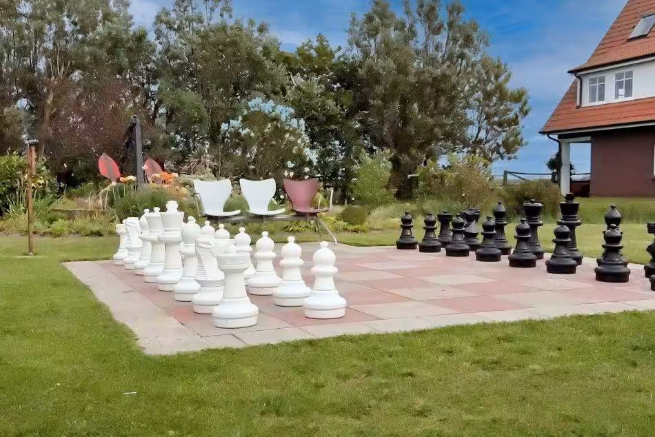 The Large Garden Chessboard Is Surrounded By Lush Green Lawns And Colorful Flower Beds. The Life-size Chess Pieces Are Placed On The Tiles Of The Chessboard, Waiting To Be Moved In An Exciting Game.