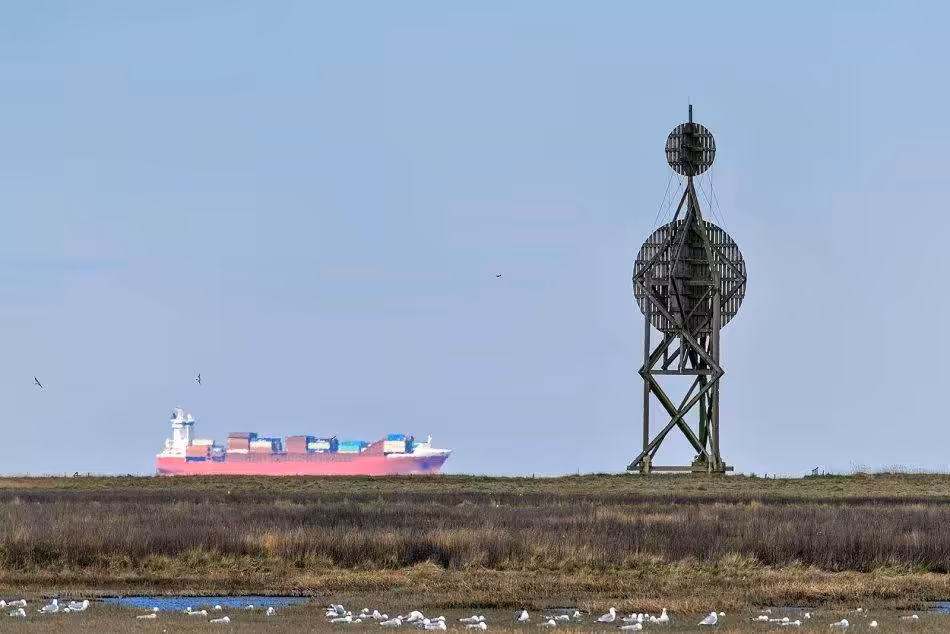 The Historic Ostbake On The Island Of Neuwerk Serves As An Impressive Backdrop While A Gigantic Container Ship Passes In The Distance, Highlighting The Contrast Between The Past And Present Of Seafaring.